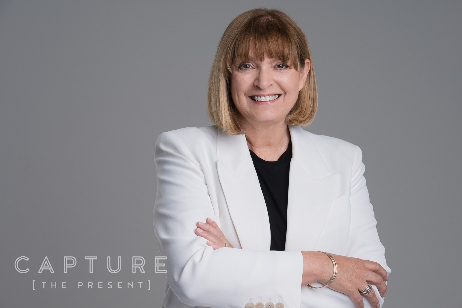 Woman with short blonde hair in a white blazer, arms folded, smiling