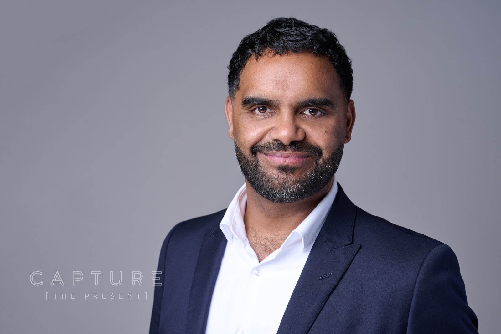 Man in a navy suit smiling against a grey studio backdrop