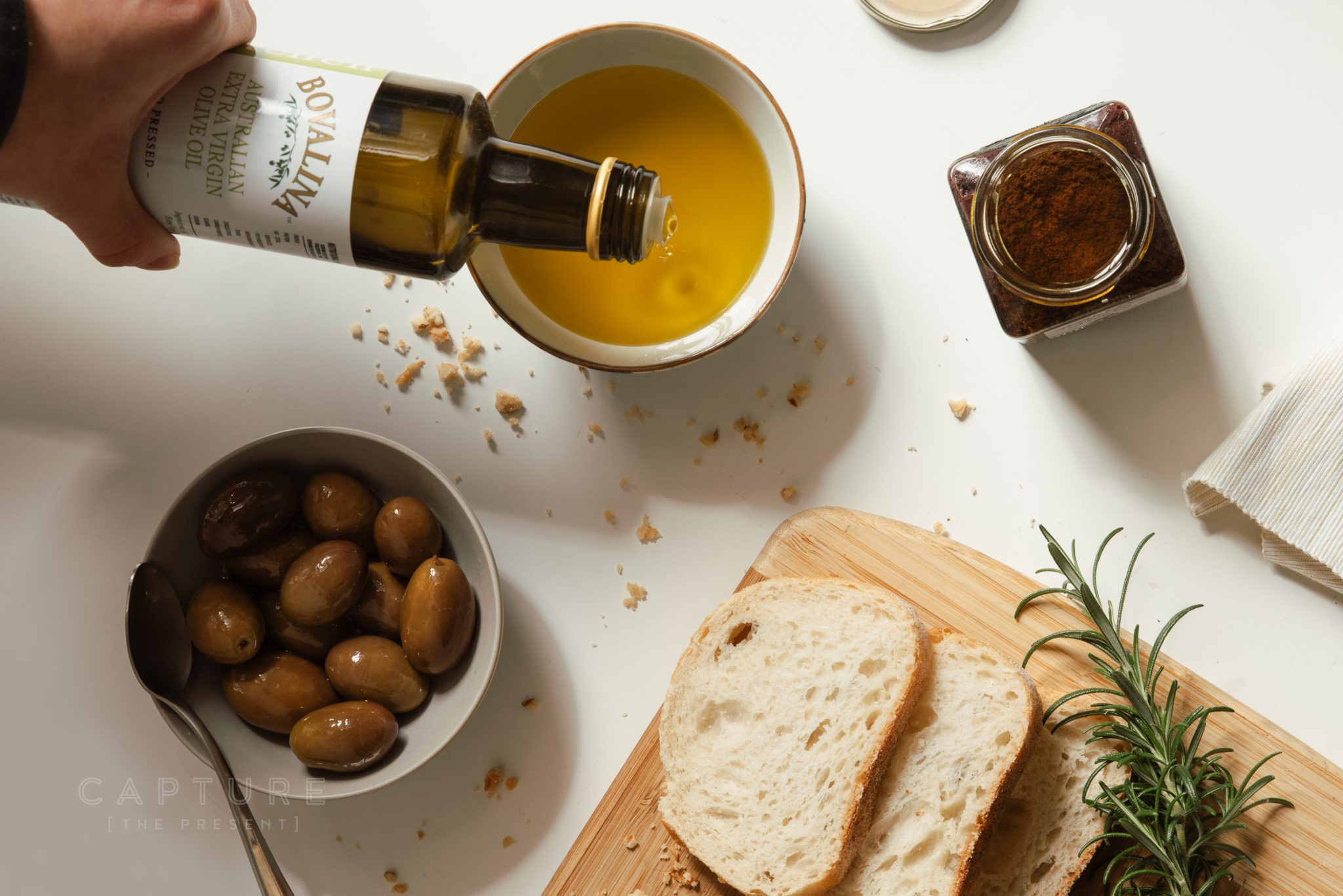 Hand pouring Bovalina olive oil into a dish beside bread, olives and rosemary