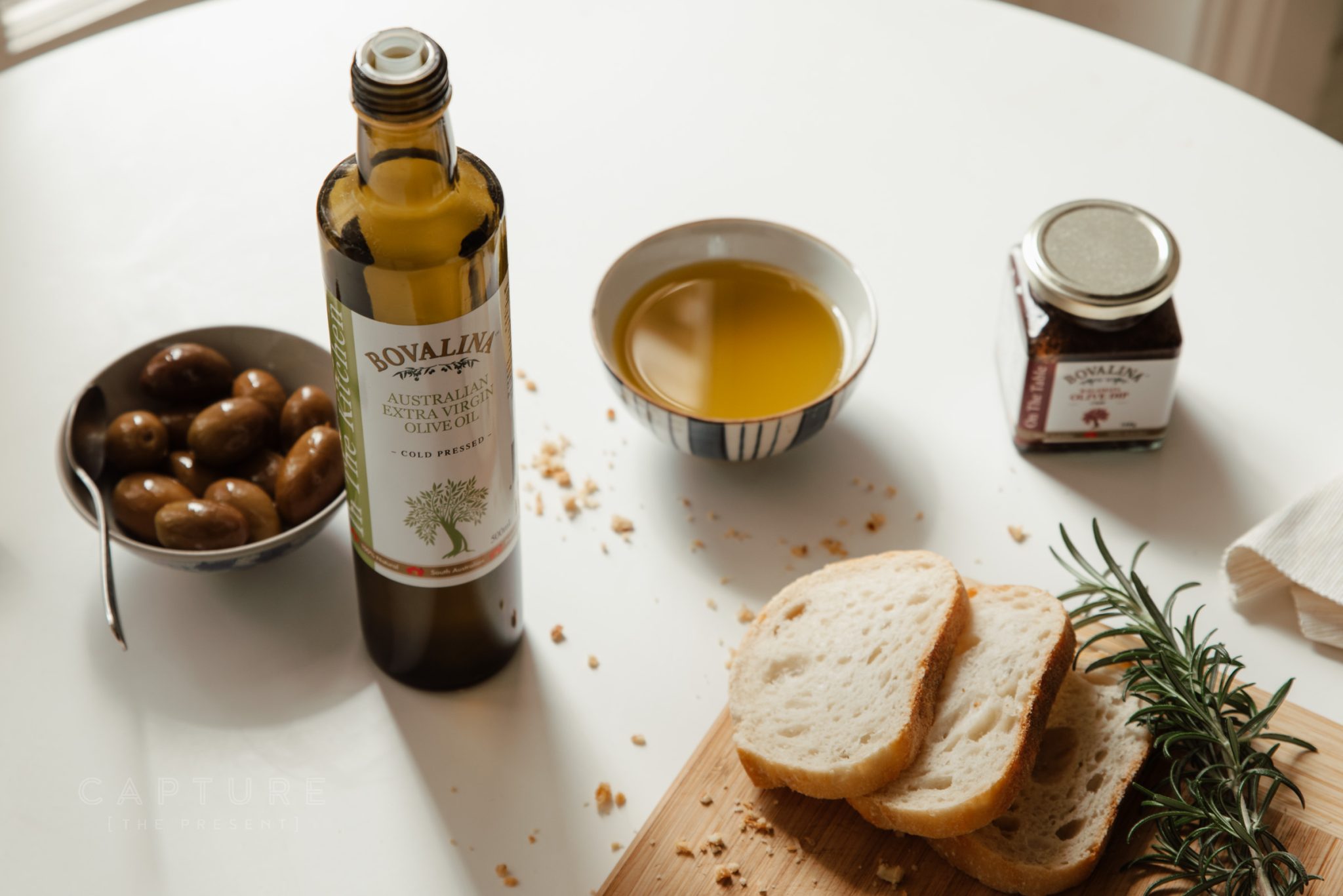 Bovalina extra virgin olive oil with olives, bread and rosemary on a styled table