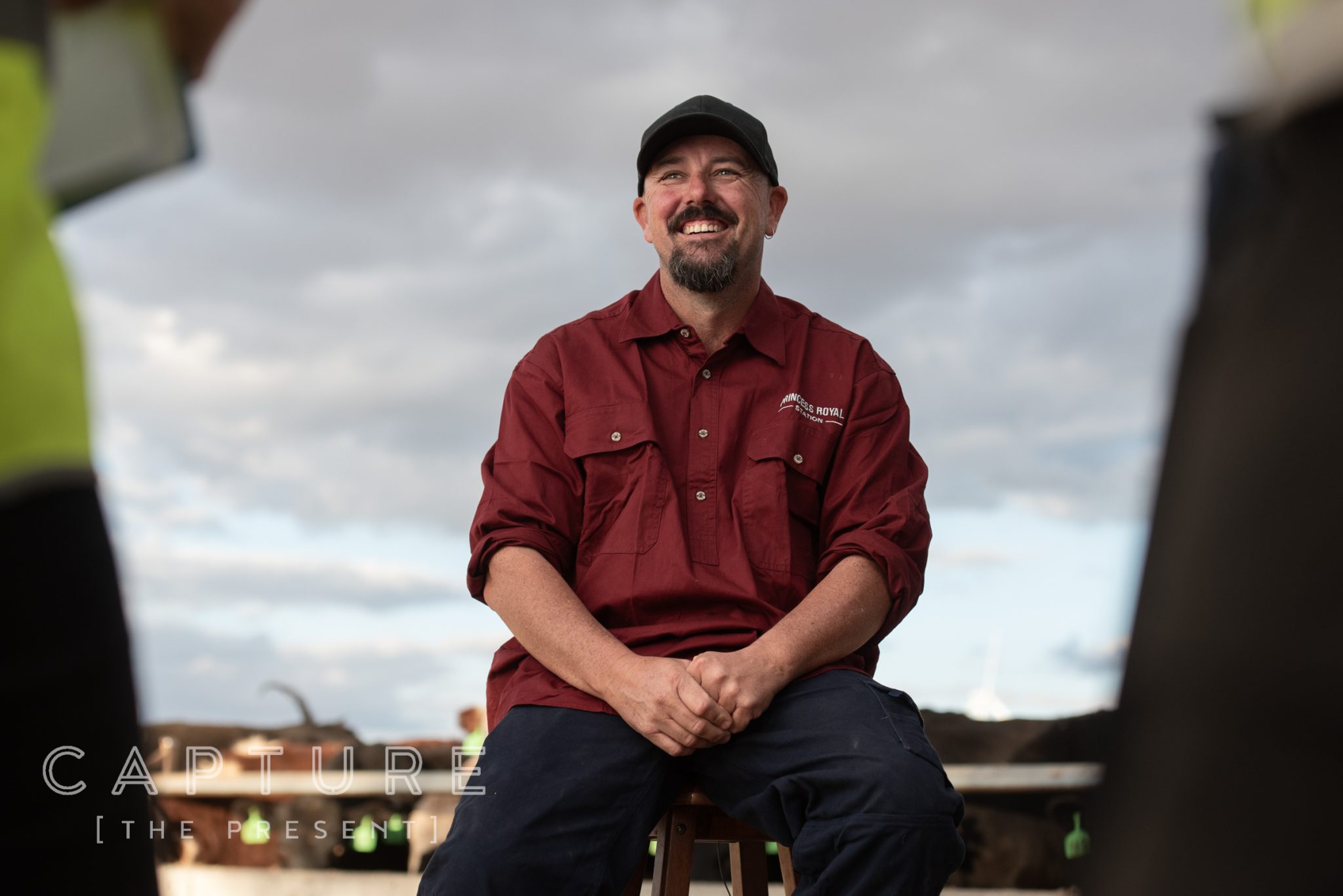 Farmer in a Princess Royal Station shirt laughing, seated at the cattle yards