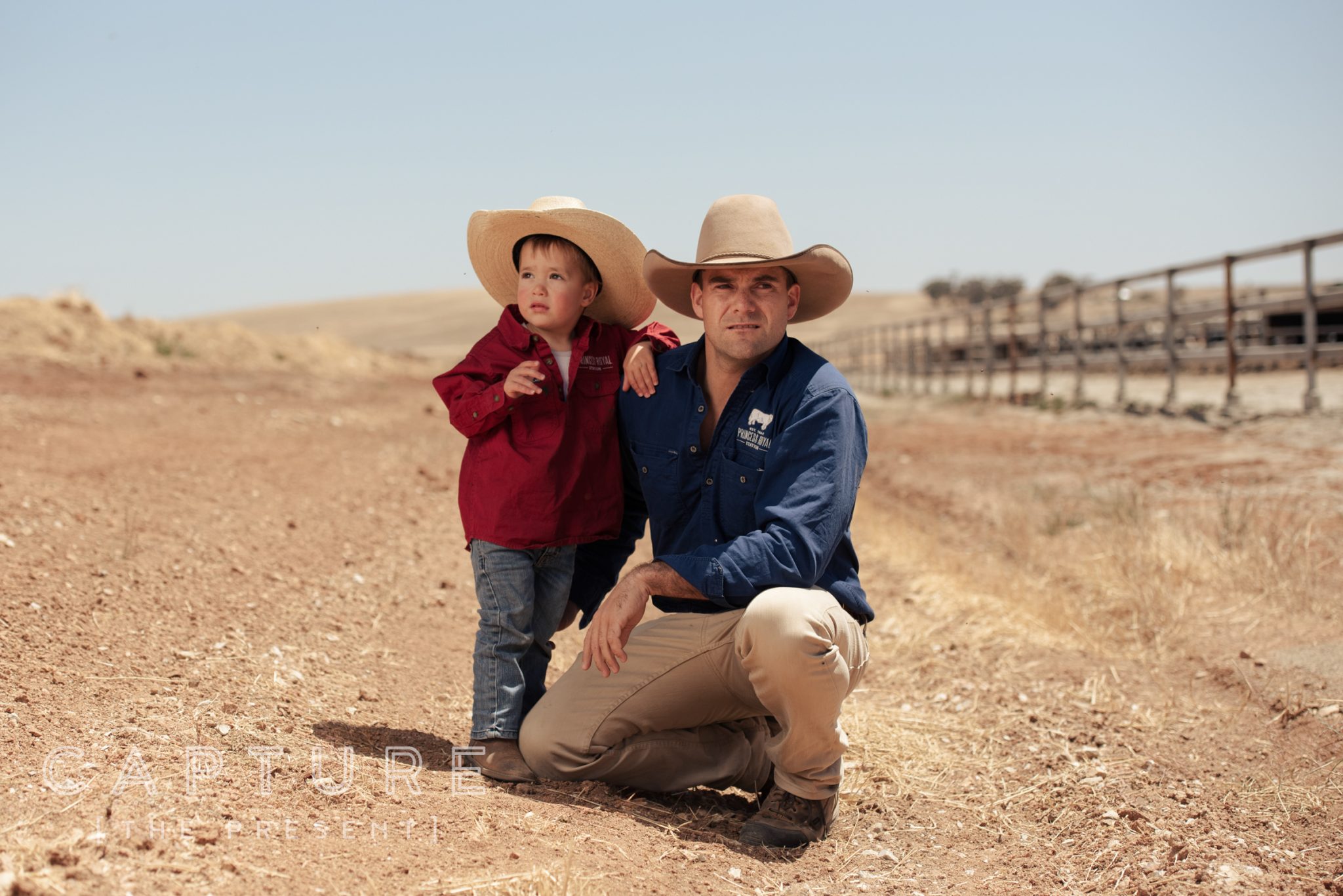 Farmer kneeling beside a young child in a matching wide brim hat at the feedlot